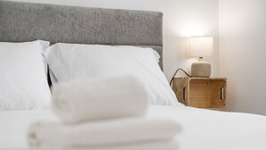 A neatly made bed with white linens and a folded towel, a grey upholstered headboard, and a bedside table with a lamp and a wooden box. - GuestReady - Oporto Old Town House 3 (Porto)