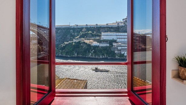 A view from a room with a red-framed window overlooking a coastal area with buildings, a bridge, and a boat on the water. - GuestReady - Oporto Old Town House 3 (Porto)