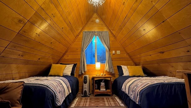 A cozy attic bedroom with two beds, a central table, and a window with curtains. - Cozy A-frame Cabin with WiFi and AC in charming Antelope (Antelope)
