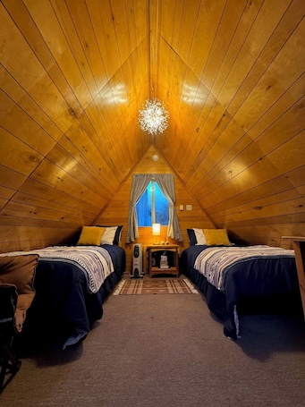 A cozy attic bedroom with two beds, a central table, and a window with curtains.