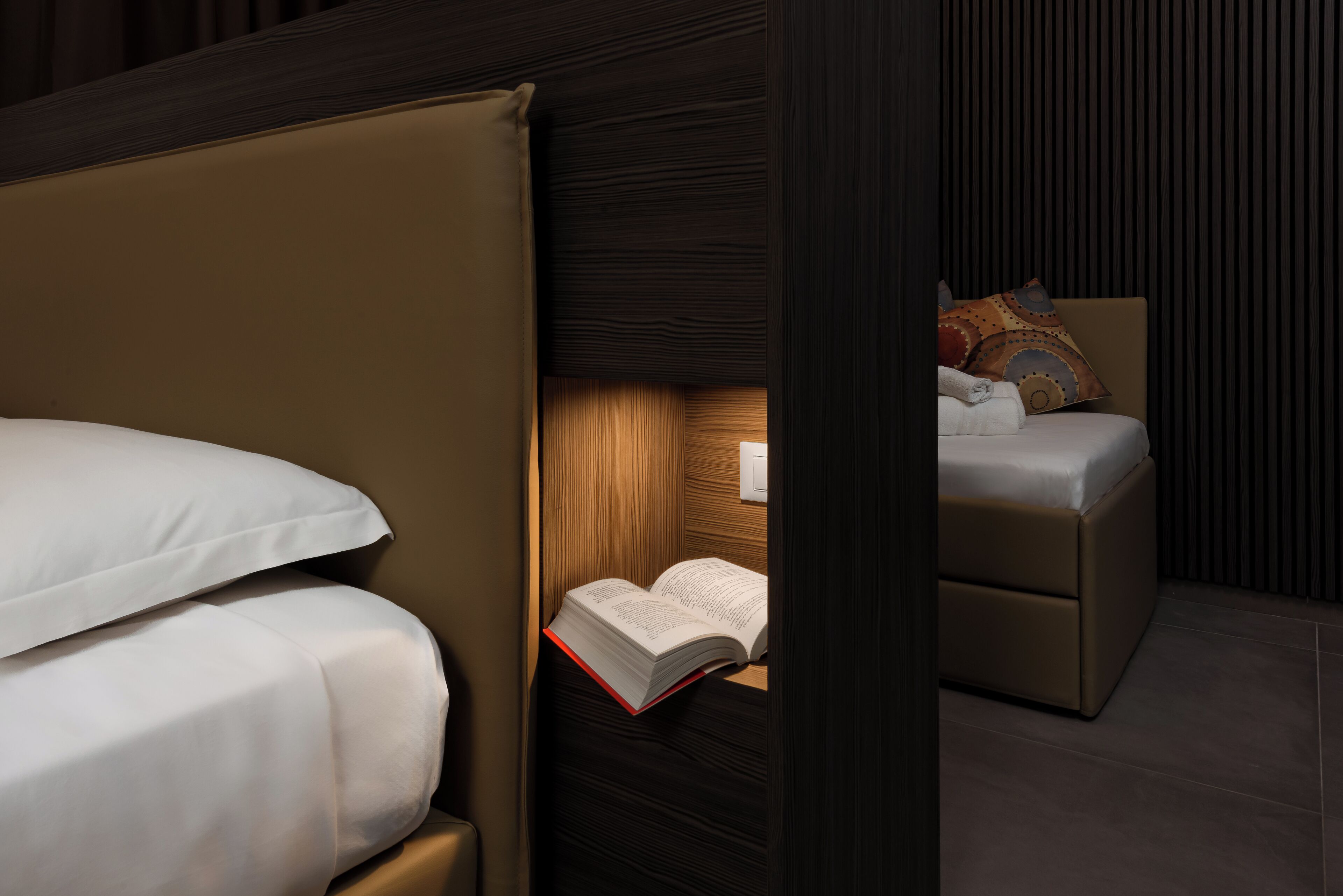 A bedroom with a bed, a reading nook with a chair and a book, and a small table with a lamp.