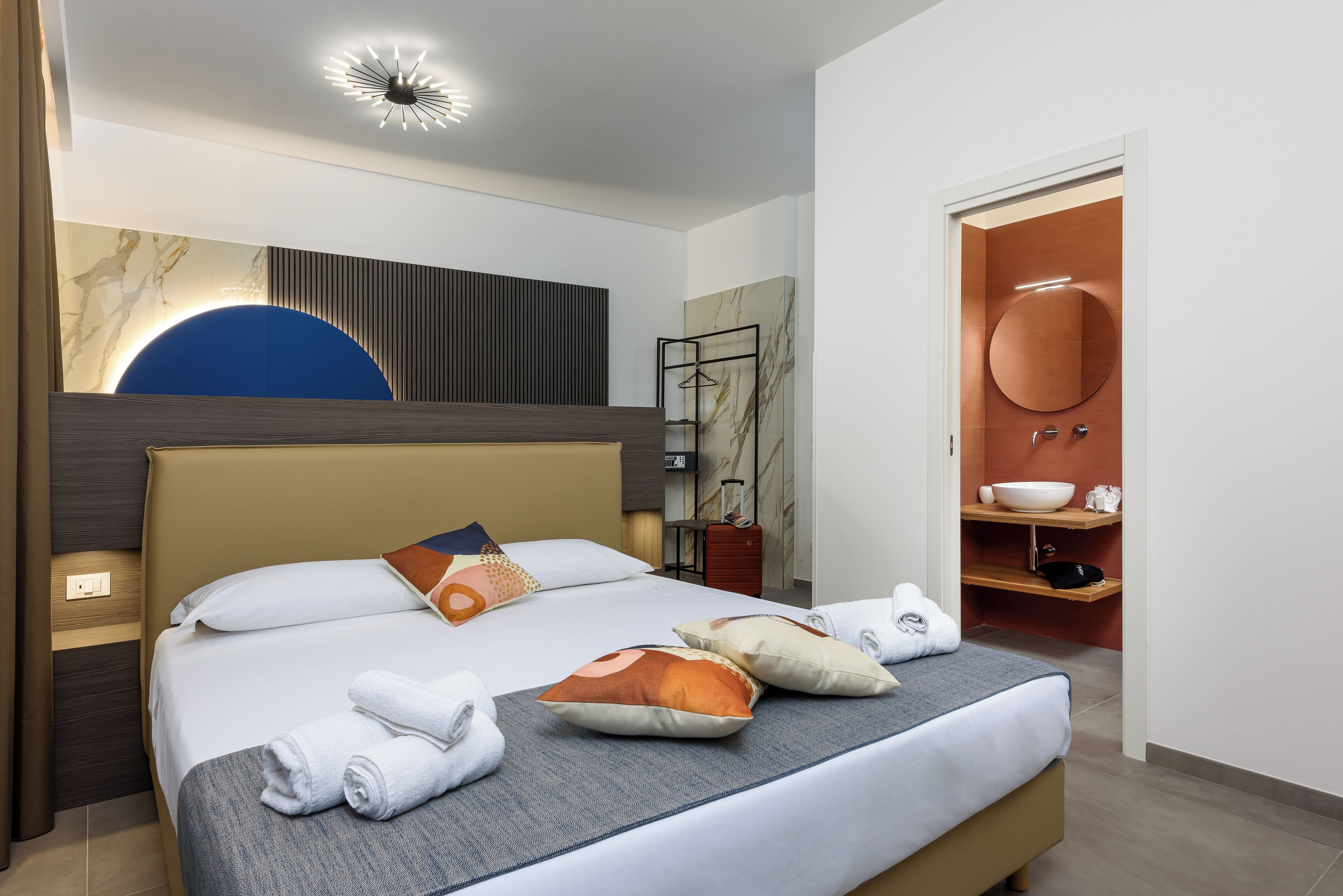 A hotel room with a large bed, a bedside table, a wardrobe, a bathroom with a sink and mirror, and a modern ceiling light.