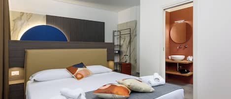A hotel room with a large bed, a bedside table, a wardrobe, a bathroom with a sink and mirror, and a modern ceiling light.