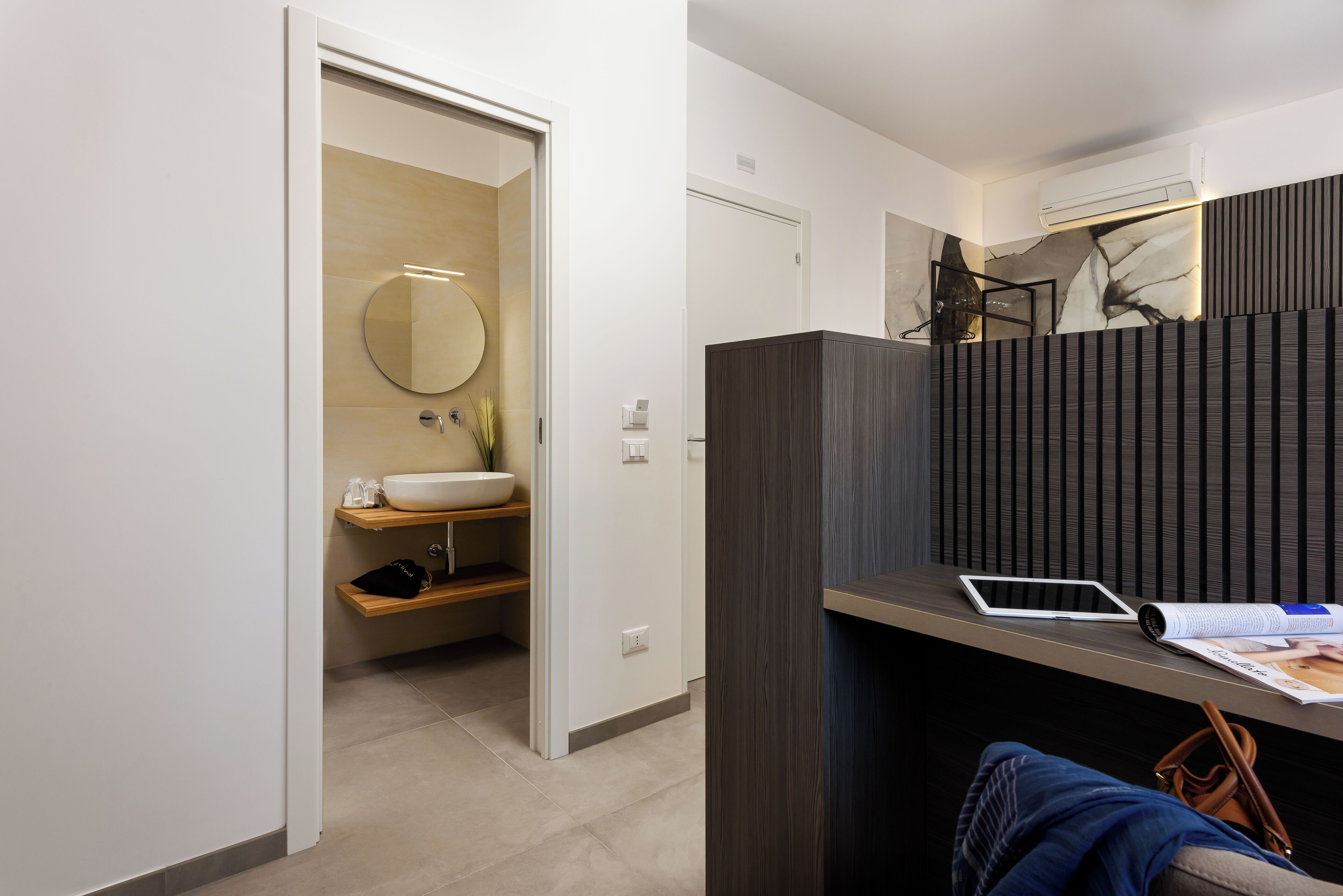 A modern bathroom with a round mirror, a sink, and a vanity. There is a tablet and a magazine on a shelf.