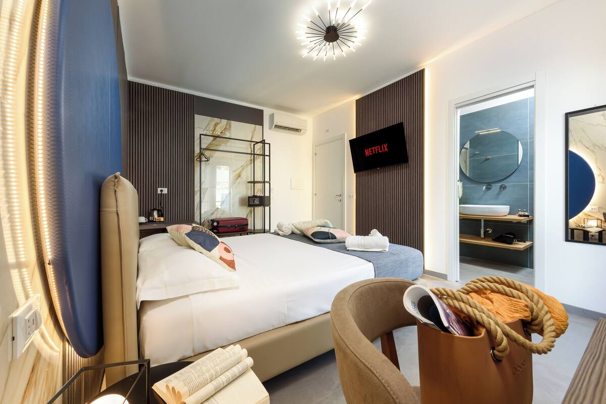 A modern hotel room with a large bed, a TV, a bathroom with a mirror and sink, and a chair with a bag.
