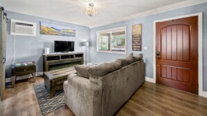 Ein Wohnzimmer mit grauem Sofa, einer Holz-Unterhaltungsecke und einem Fernseher. Eine Tür befindet sich rechts, und ein Fenster mit Jalousien ist vorhanden.