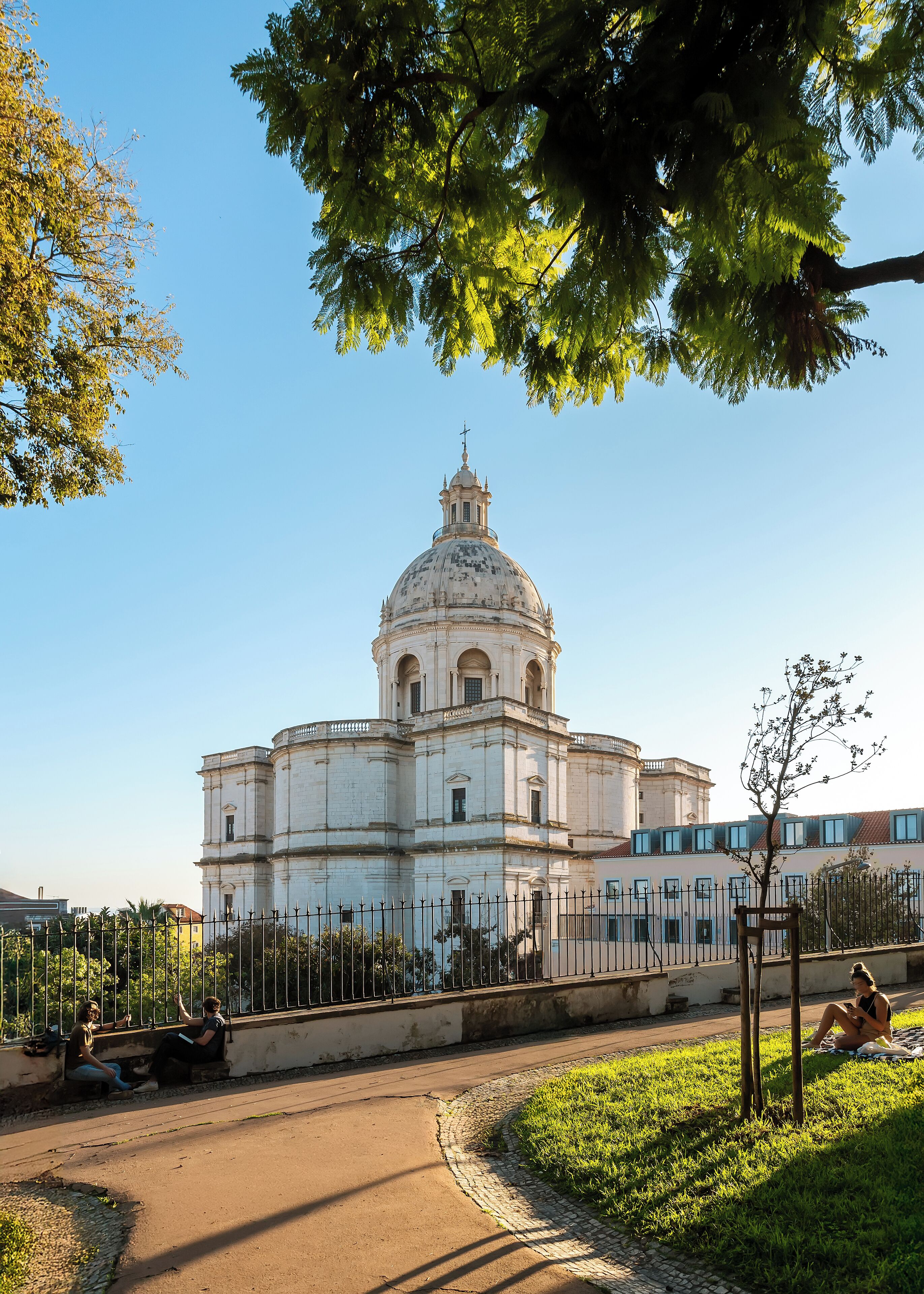 Un edificio grande e histórico con una cúpula destacada y un campanario, rodeado de una cerca y follaje.