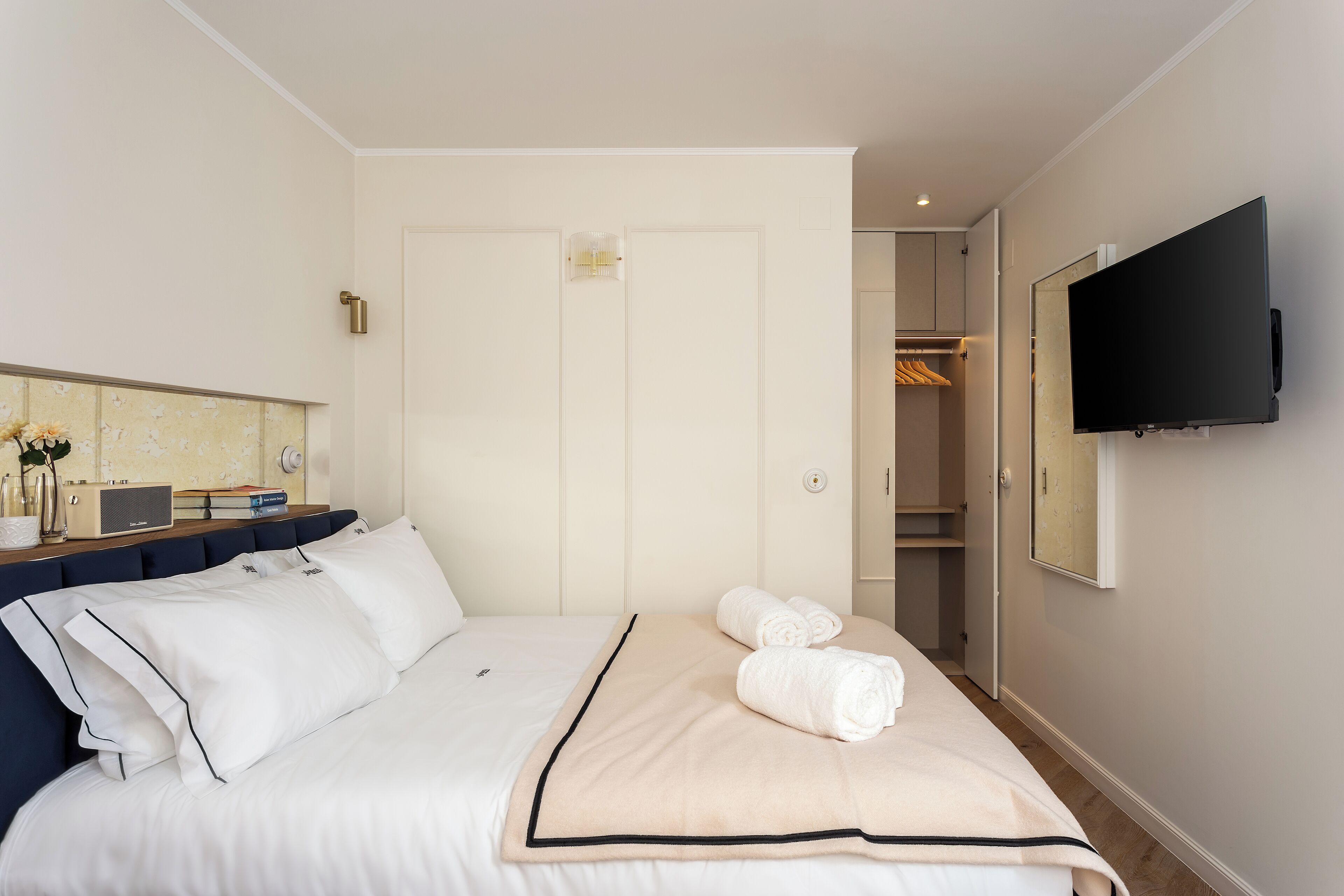 A neatly made bed with white bedding and a beige throw blanket, a bedside table with a lamp, and a TV mounted on the wall.