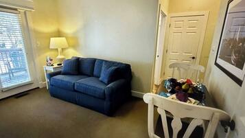Une pièce chaleureuse avec un canapé bleu, une petite table sur laquelle se trouve une lampe, et un fauteuil avec un panier de fruits.