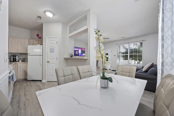 A modern kitchen with white cabinets, a white refrigerator, and a dining area with a marble table and beige chairs.