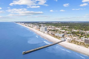 A coastal cityscape with a long pier extending into the ocean, surrounded by high-rise buildings and a beach.