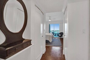 Corredor de hotel com um espelho grande, penteadeira de madeira, cama com tapete listrado e varanda com vista.
