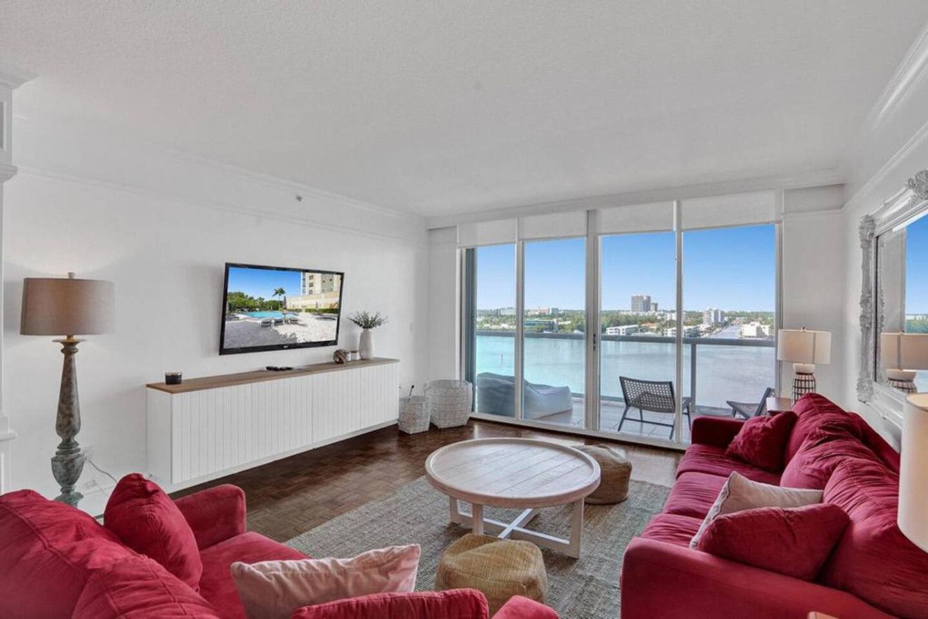 A modern living room with a large TV, red sofas, a round coffee table, and a view of a body of water through large windows.