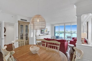 A spacious living room with a wooden dining table, red and white furniture, a large window with a view of the sea, and a hanging light fixture. - Le Petit Italie ,Water view 2/2 with pool (Sunny Isles Beach)