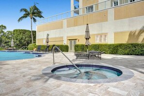 Un espace piscine avec des chaises longues et des parasols, un jacuzzi et un bâtiment doté de balcons.