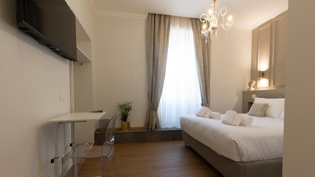 A modern bedroom with a bed, bedside table, TV, and a window with curtains. - De Vince Suites   Luxury Rooms   King Executive (Roma)