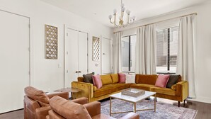 A living room with a mustard-colored sofa, a brown leather armchair, and a coffee table. There are two doors with decorative panels, a chandelier, and a window with blinds. - Stylish Pearl District Stay - Relax, Dine & Explore (San Antonio)