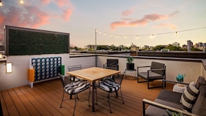 A rooftop terrace with a wooden table and chairs, string lights, a green wall, and a city view. - Stylish Pearl District Stay - Relax, Dine & Explore (San Antonio)