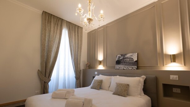 A neatly made bed with pillows and a folded towel, a chandelier, a painting of a building, and a window with curtains. - De Vince Suites   Luxury Rooms   Roma Termini   King De Luxe (Roma)