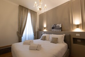 A neatly made bed with pillows and a folded towel, a chandelier, a painting of a building, and a window with curtains.