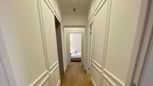 A narrow hallway with white walls and wooden flooring, leading to a bedroom with a bed and a nightstand. - De Vince Suites   Luxury Rooms   Roma Termini   King De Luxe (Roma)