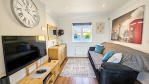 A modern living room with a large flat-screen TV, wooden furniture, a patterned rug, and a large wall clock. - Boughton 3 Bed Semi by Fortuna Property Portfolio (Gloucester)