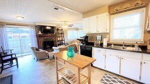 A spacious living room with a fireplace, a dining area with a wooden table, and a kitchen with white cabinets and a window.
