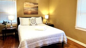 A bedroom with a bed, two bedside tables with lamps, a painting on the wall, and a window with blinds.