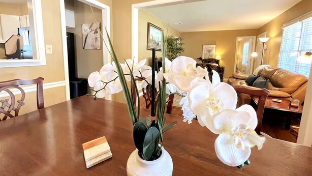 A dining area with a wooden table, white orchid flowers, and a vase. There is a brown leather couch and a framed picture on the wall in the background.