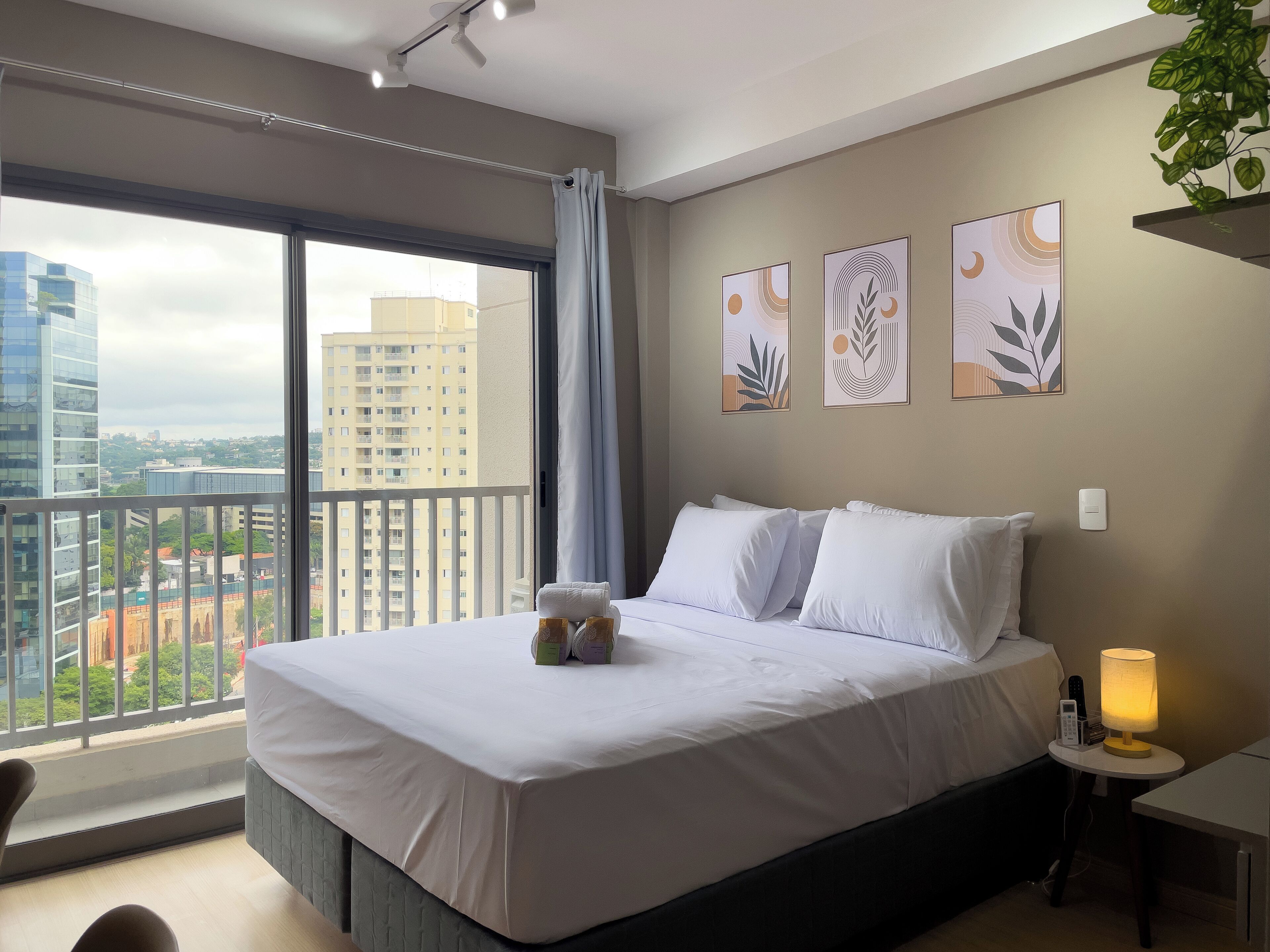 A modern hotel room with a large bed, a bedside table with a lamp, and a view of the city through a balcony.