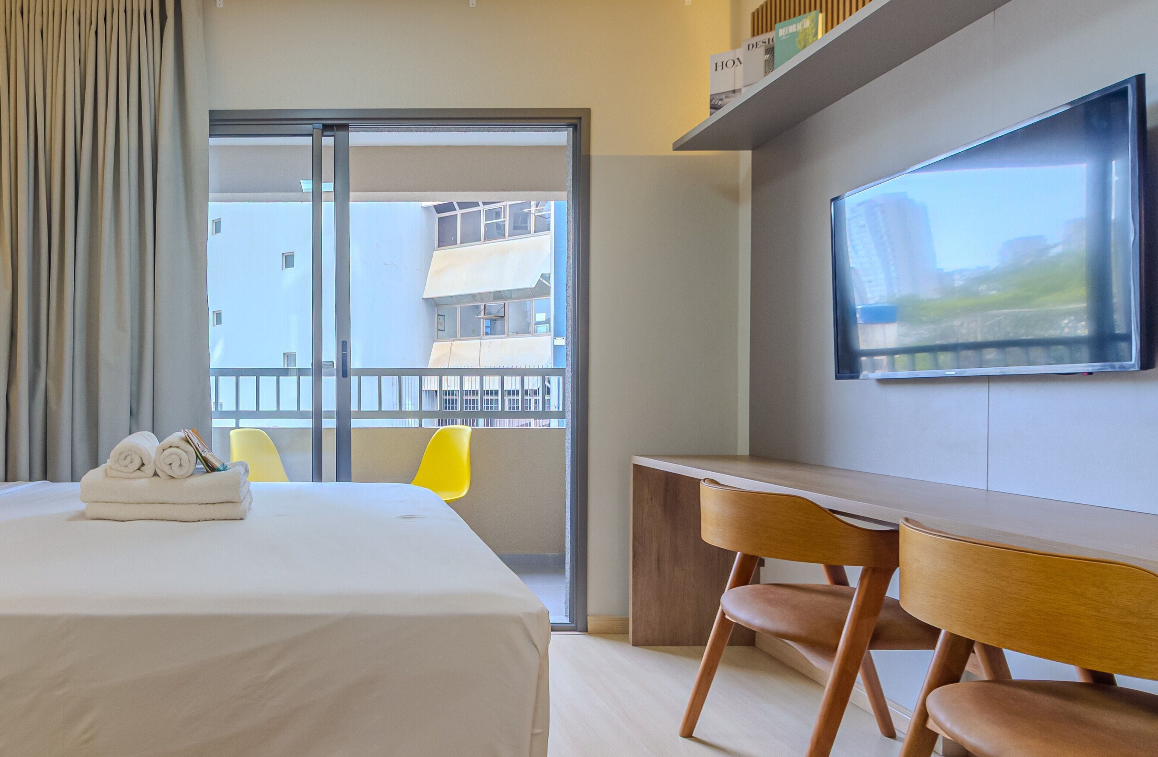 Quarto de hotel com cama, secretária, duas cadeiras, televisão e varanda com vista para edifícios.