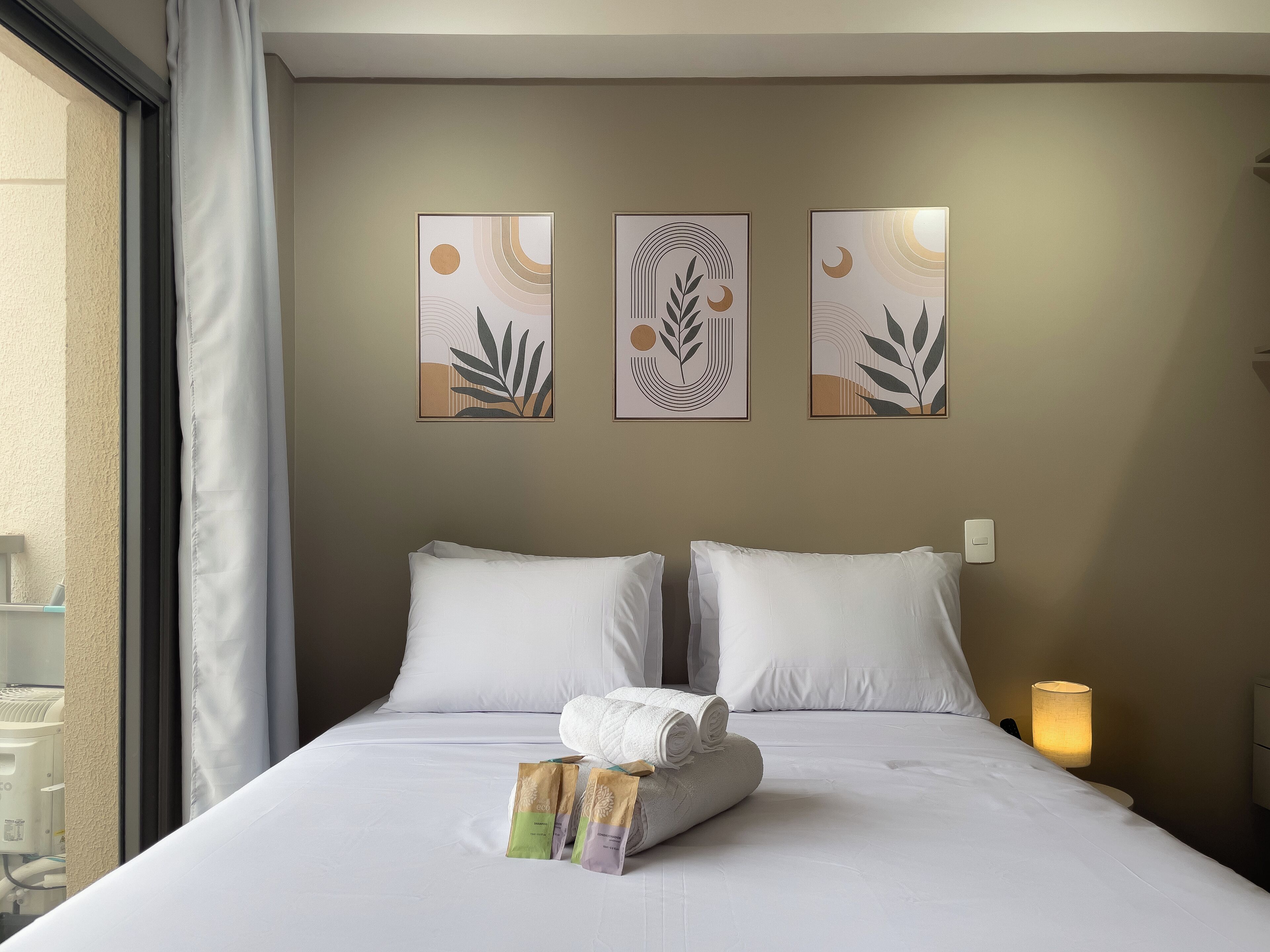 Une chambre d’hôtel avec un lit, des oreillers blancs, une serviette roulée et trois trousses de toilette. Trois œuvres d’art encadrées sont accrochées au mur au-dessus du lit.