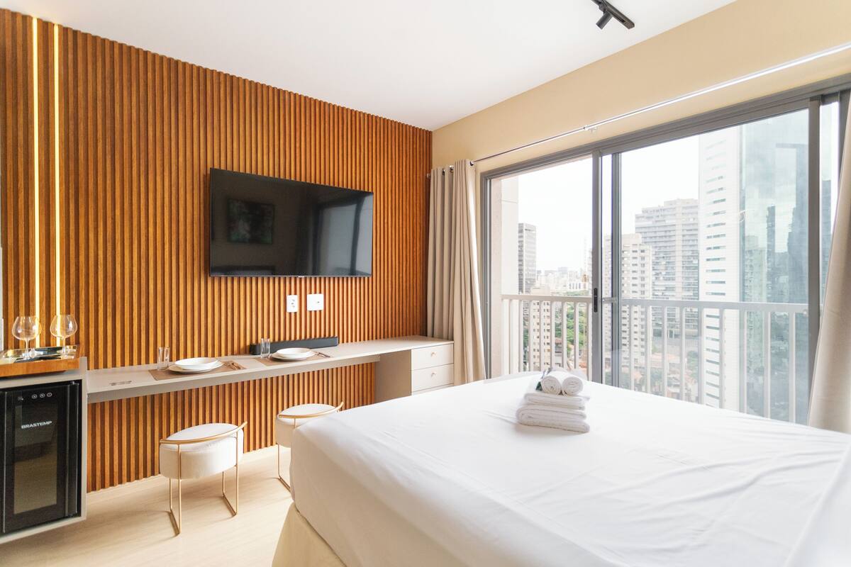 A modern hotel room with a wooden accent wall, a flat-screen TV, a balcony with a city view, a bed with white linens, and a minibar.