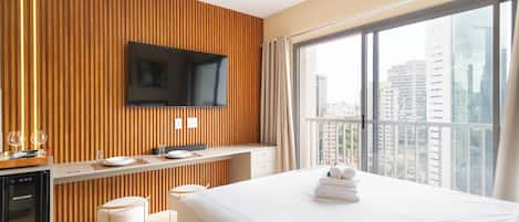 A modern hotel room with a wooden accent wall, a flat-screen TV, a balcony with a city view, a bed with white linens, and a minibar.