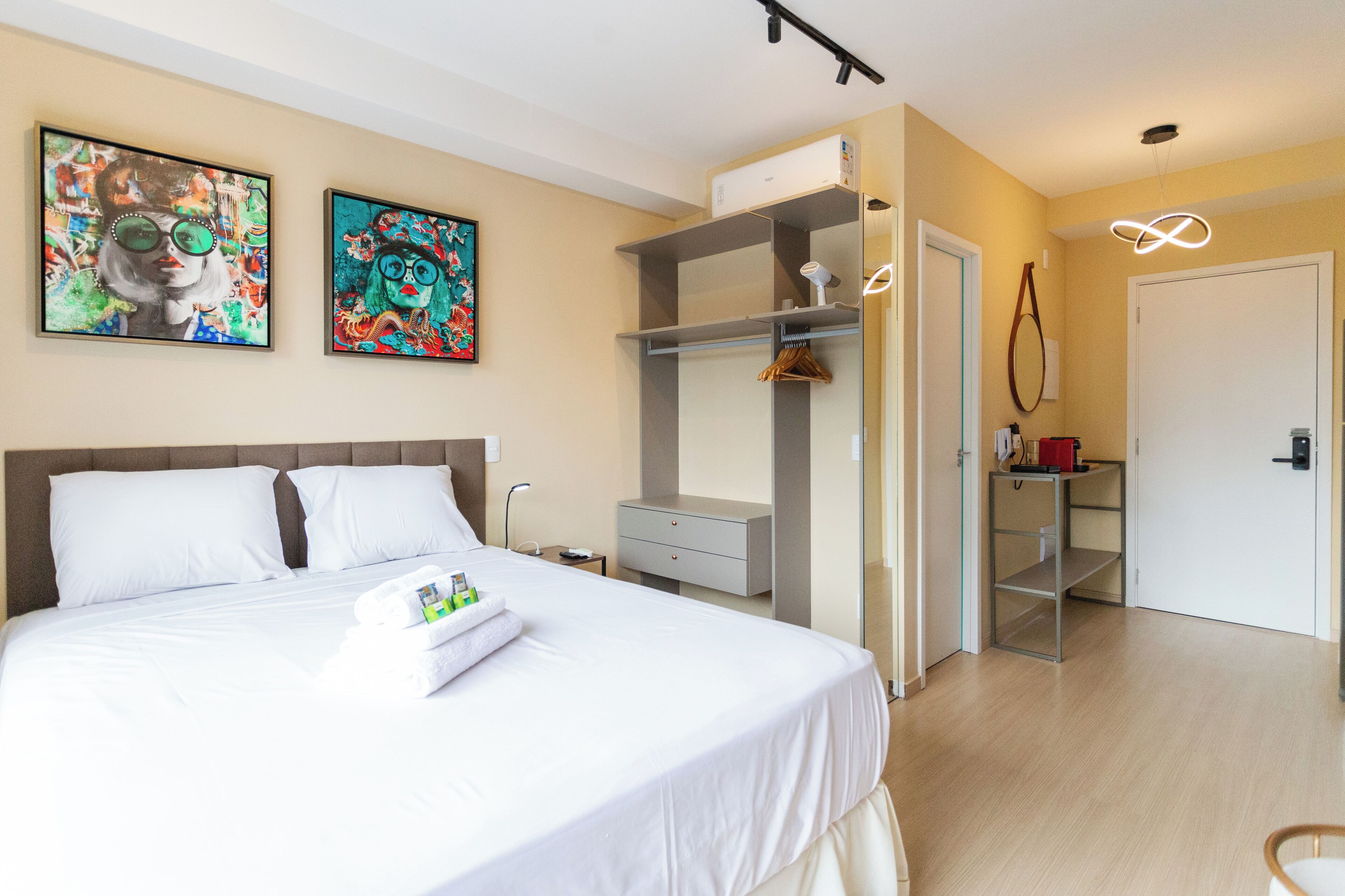 A hotel room with a bed, two paintings on the wall, a wardrobe, a desk with a lamp, and a door.