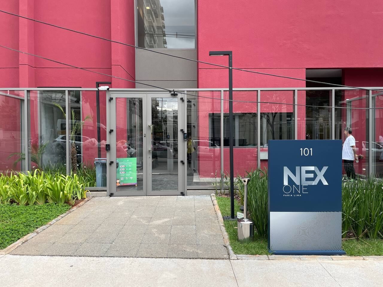 En modern byggnadsentré med en glasdörr, en skylt med texten ”101 NEX ONE” och en asfalterad gångväg som leder fram till entrén.