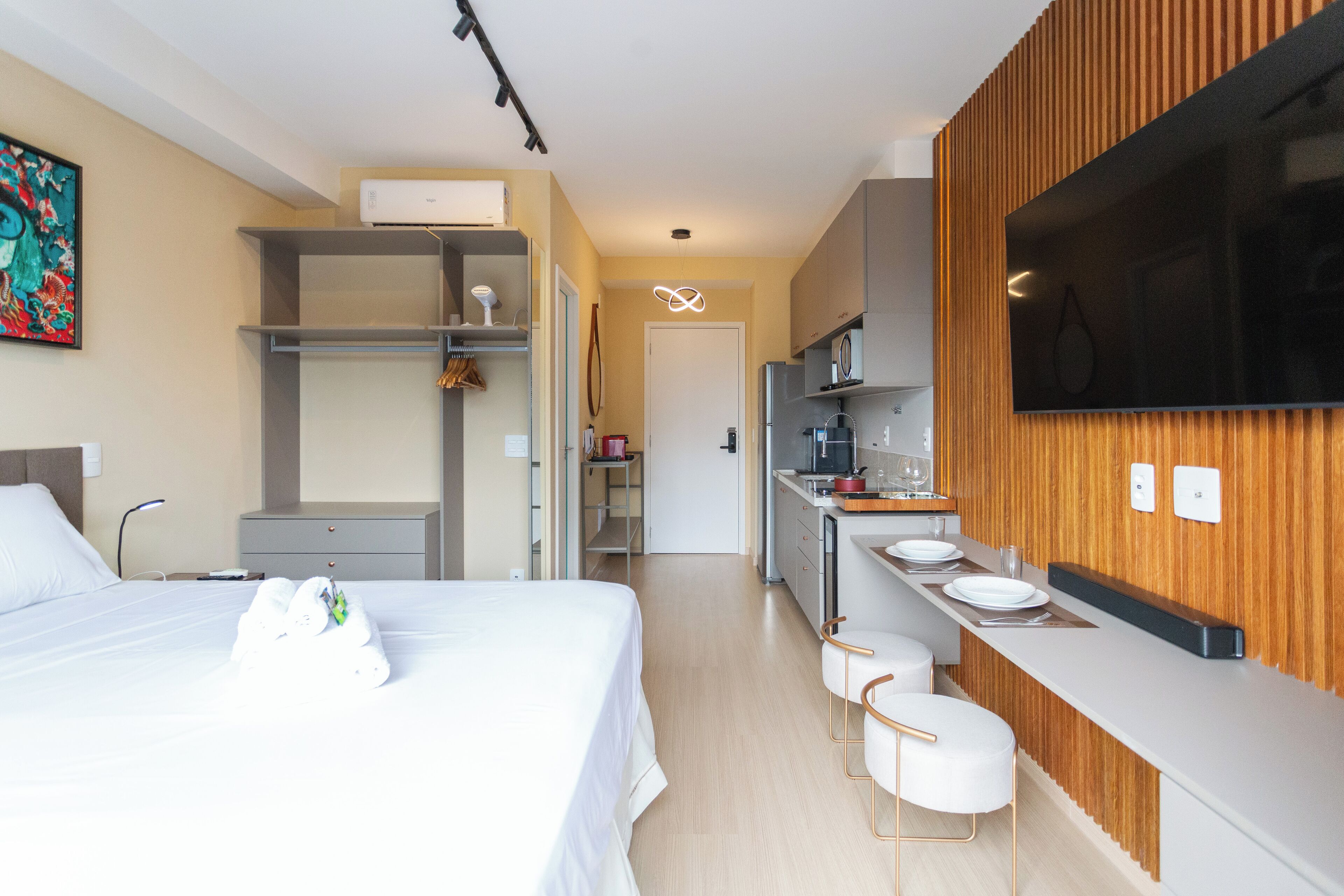 A modern hotel room with a bed, a desk, a kitchenette, and a TV.