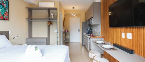 A modern hotel room with a bed, a desk, a kitchenette, and a TV.