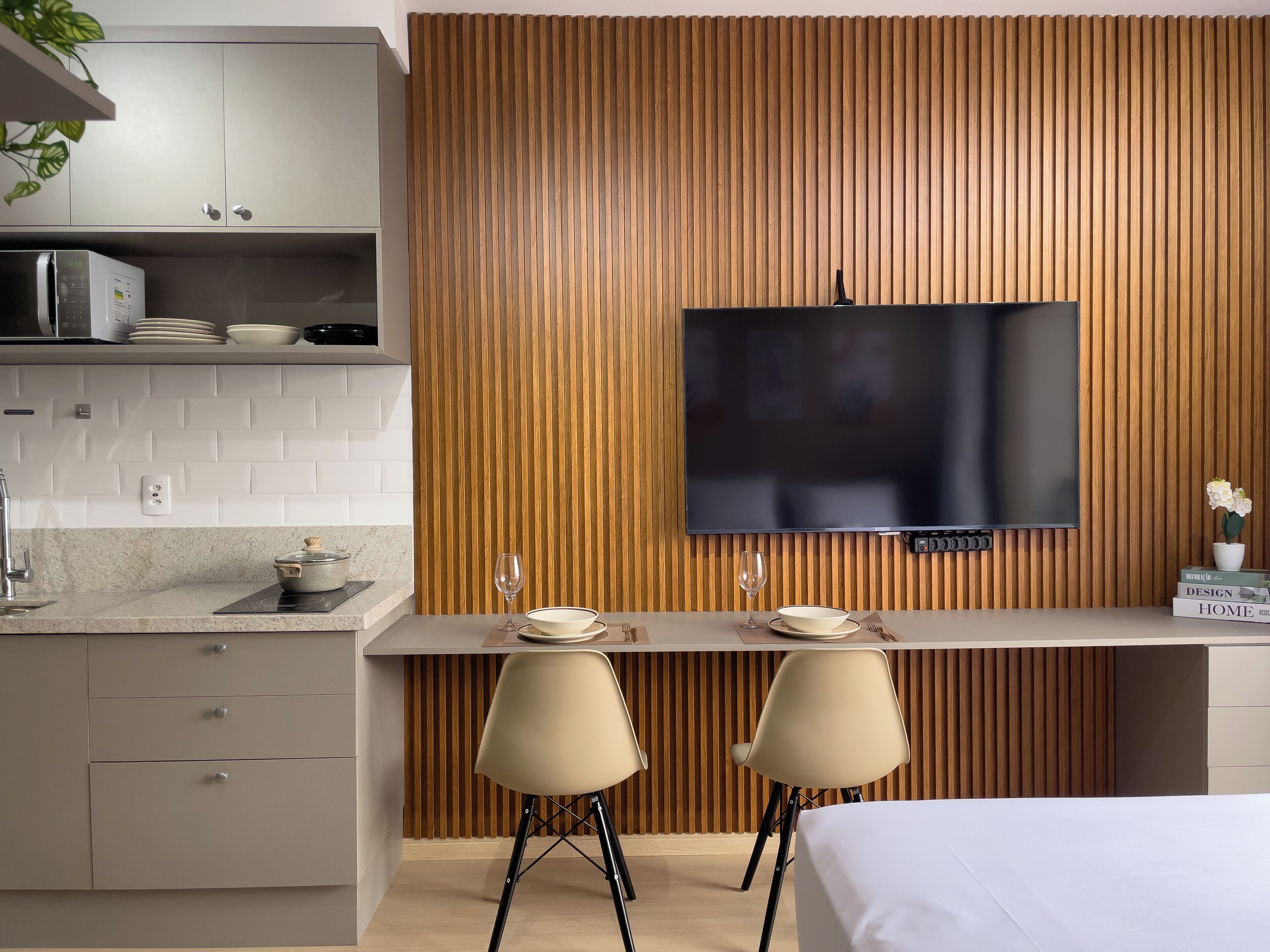 A modern kitchen with a built-in microwave, a wooden panel wall, a flat-screen TV, and a dining area with two chairs and a table.
