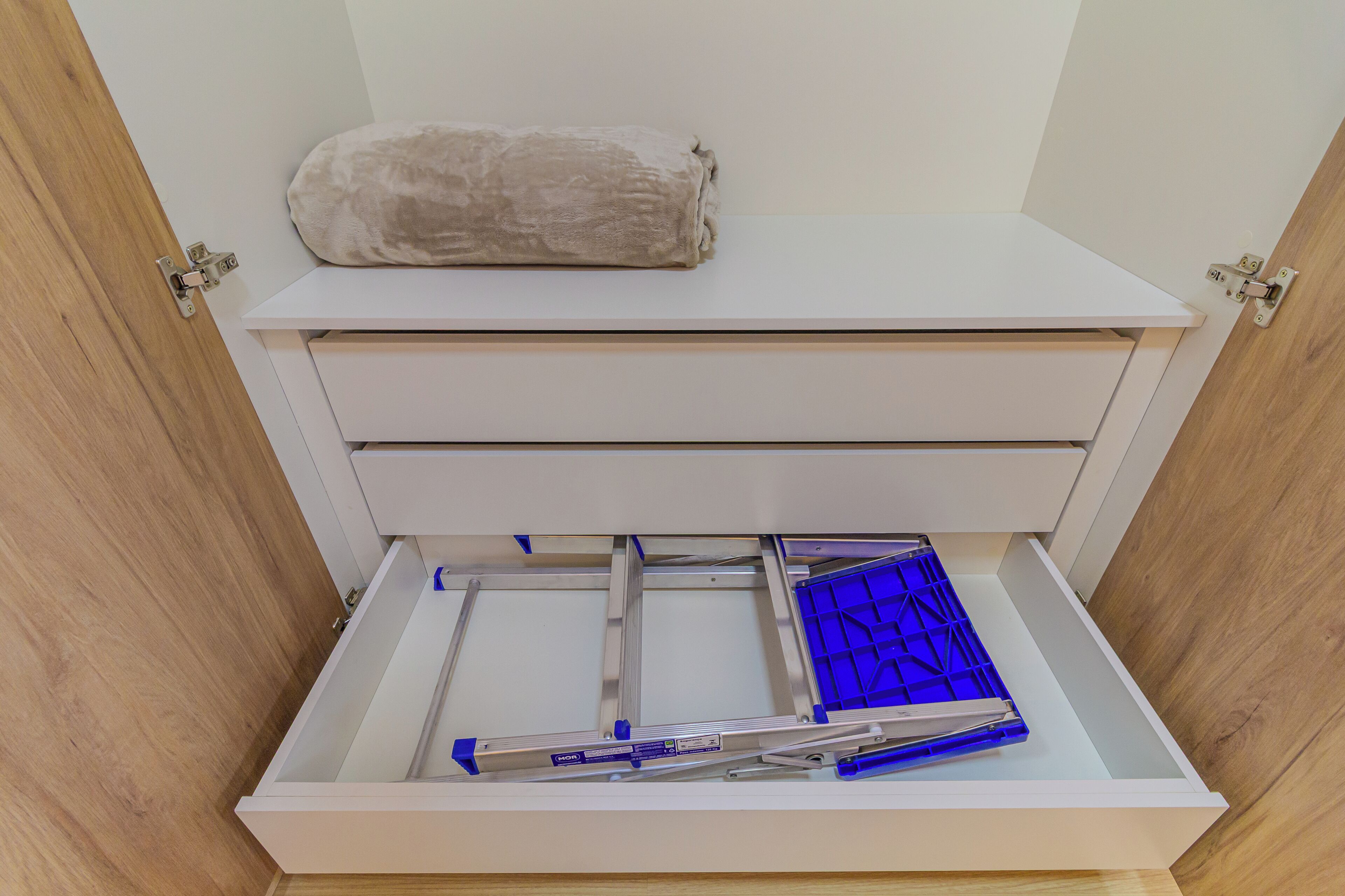 Un mobile contenitore bianco con un inserto a griglia blu, un cuscino beige sopra e un pavimento in legno.