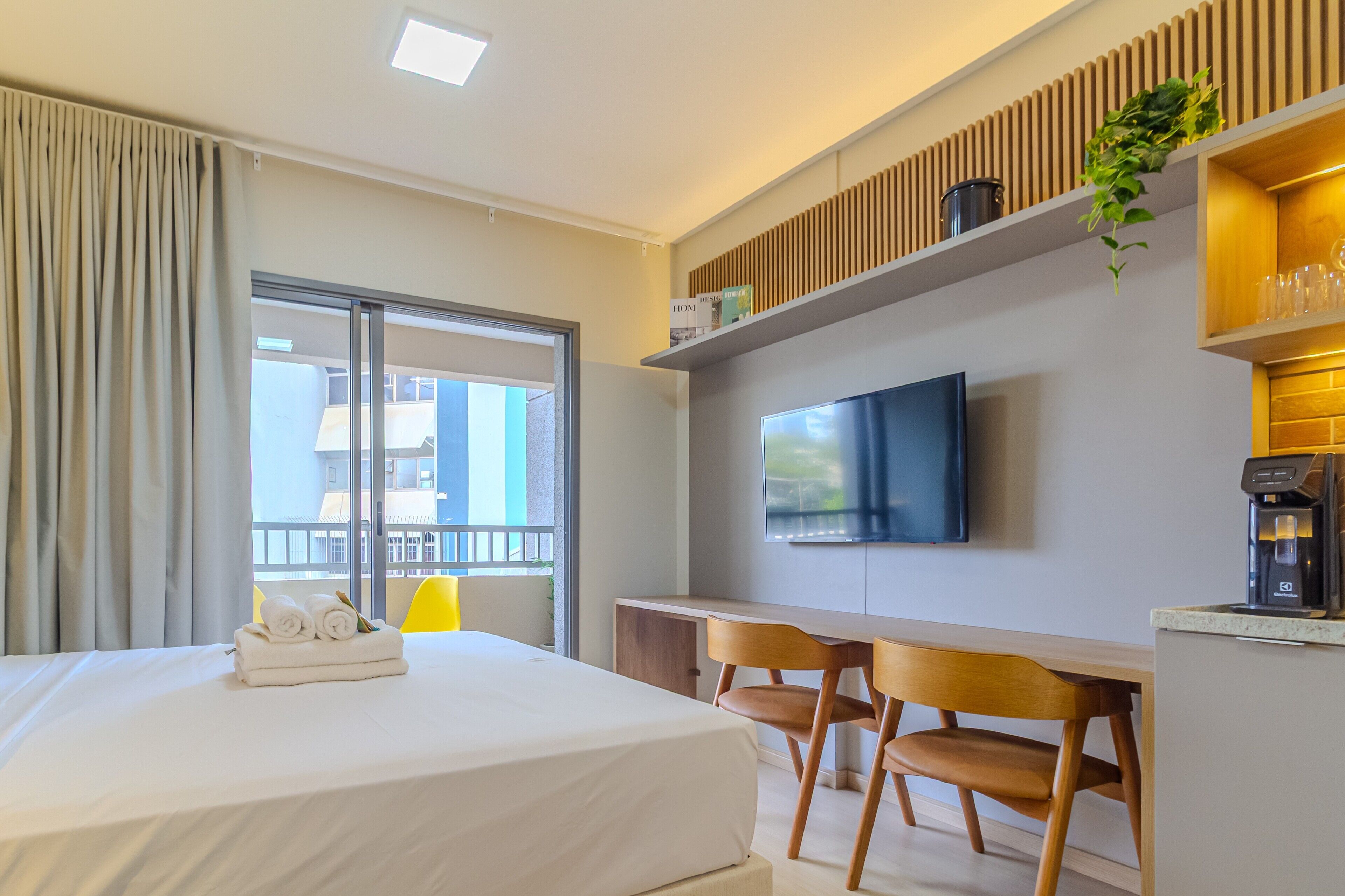 A hotel room with a bed, a desk, two chairs, a TV, a coffee machine, and a window with curtains.