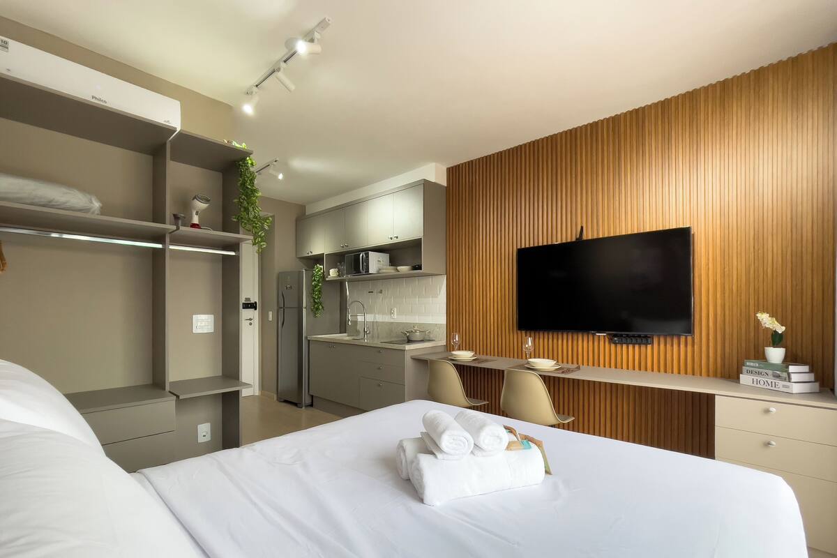 A modern hotel room with a bed, a kitchenette, a TV, and a wooden accent wall.