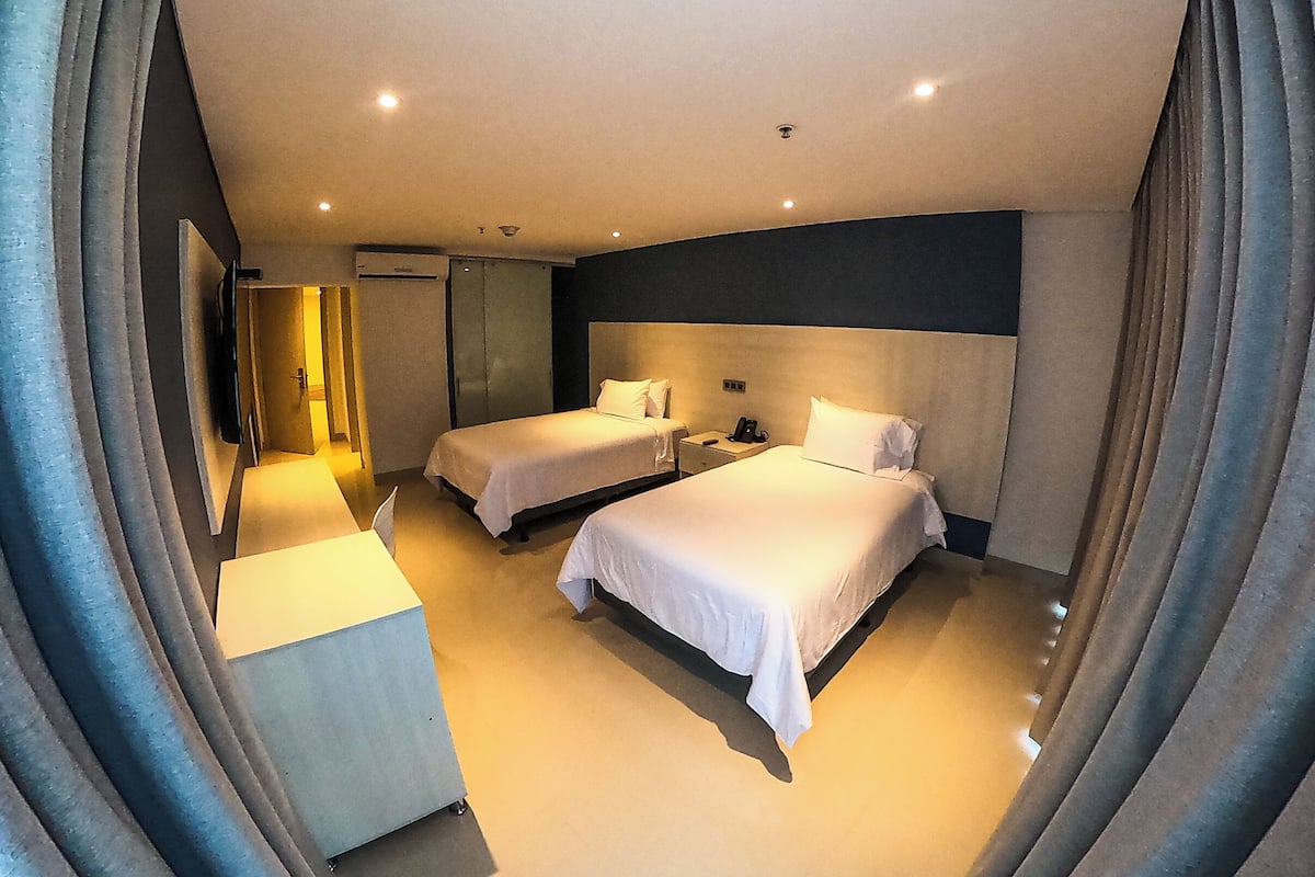A hotel room with two beds, a bedside table with a lamp, a flat-screen TV mounted on the wall, and a wardrobe.