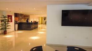 Zona de recepción de un hotel, con un mostrador, un televisor de pantalla plana grande en la pared y una planta en maceta.