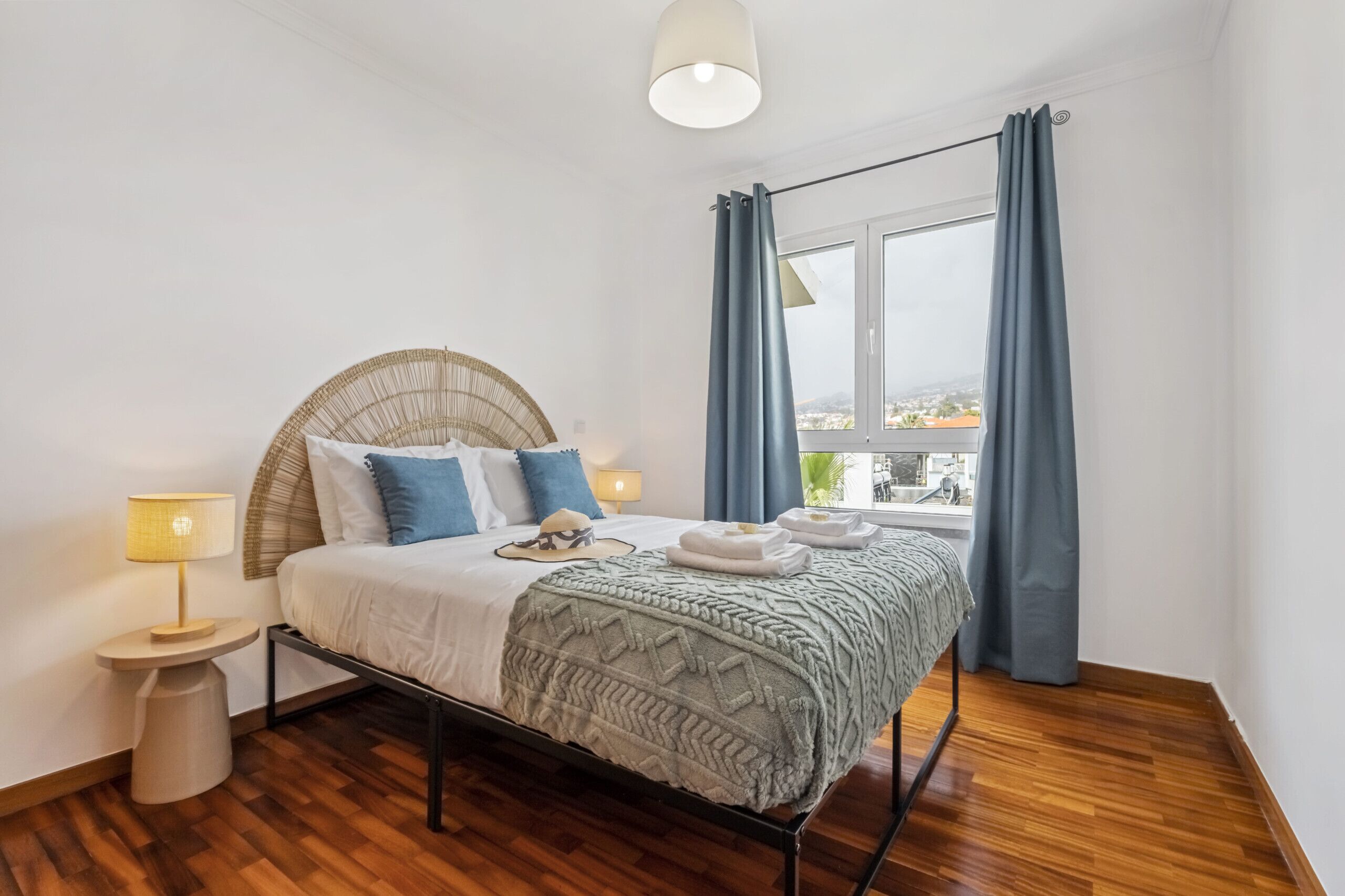 Un dormitorio ordenado con cama, lámparas de noche, una ventana con cortinas y vistas al exterior.
