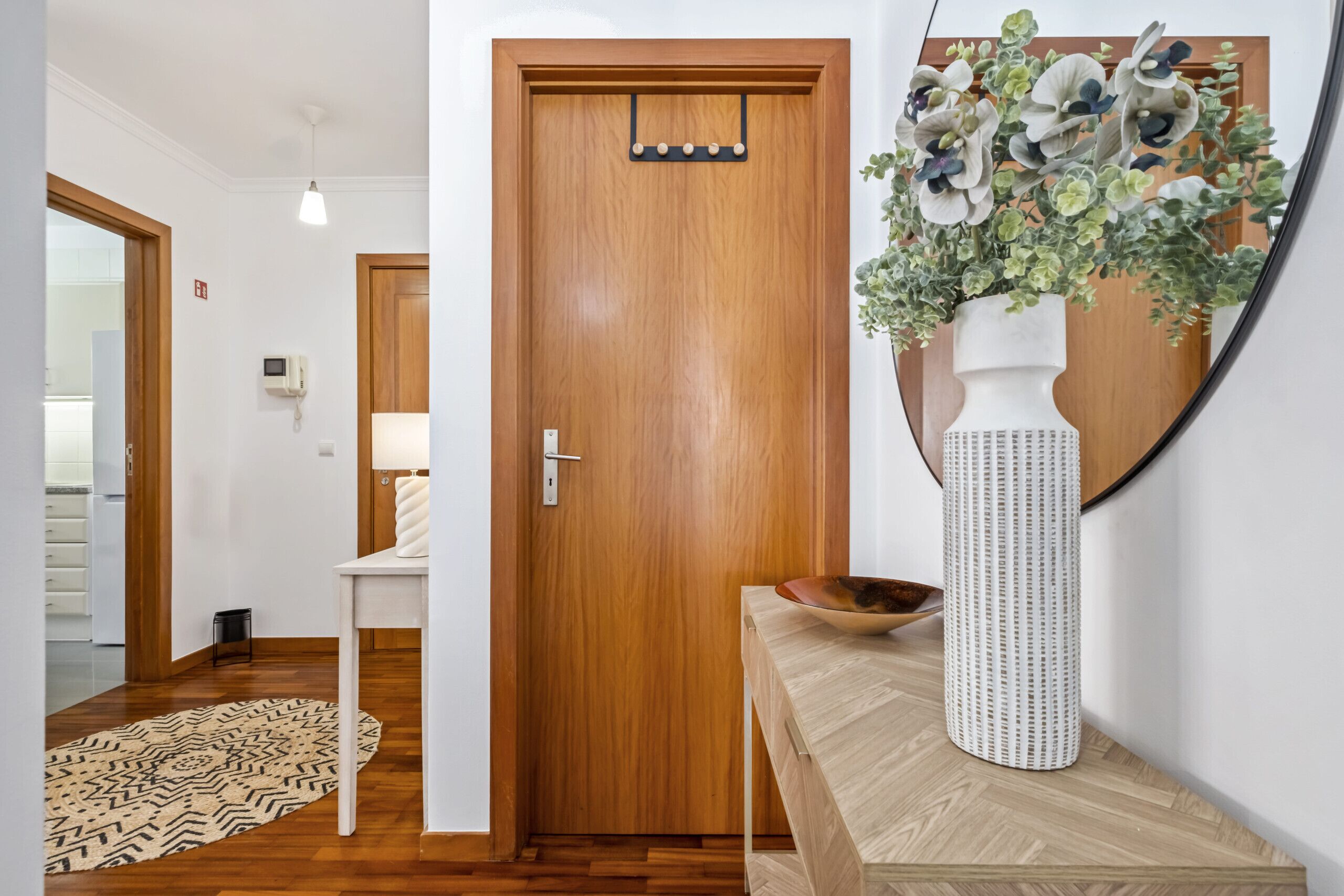 A hallway with a wooden door, a white console table, a patterned rug, and a mirror with a vase of flowers.