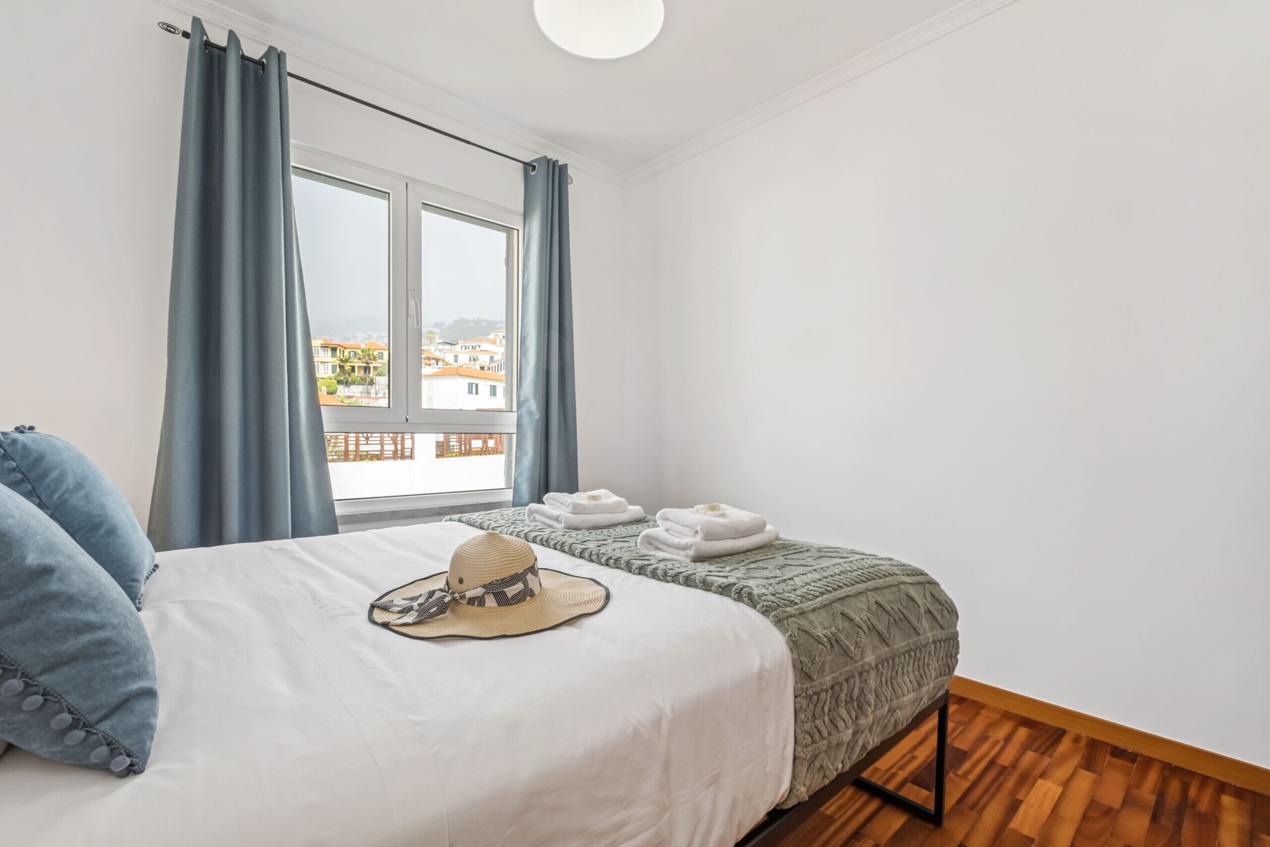 A bedroom with a bed, two bedside tables, a window with curtains, and a view of buildings outside.