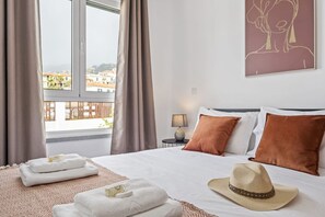 Une chambre d’hôtel avec un lit, des oreillers, un chapeau, des serviettes, une lampe et une vue sur les bâtiments par la fenêtre.