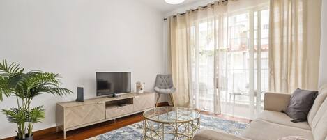 A modern living room with a sofa, a glass coffee table, a TV on a wooden cabinet, a potted plant, and a large window with curtains.