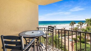 A balcony with a table and chairs, overlooking a beach with palm trees and a pool area.